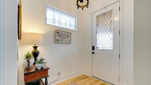 Un couloir doté d’un plancher en bois, d’une porte blanche ornée d’un panneau de verre décoratif, d’une carte accrochée au mur, d’une lampe et d’une petite table sur laquelle sont disposés des objets décoratifs.