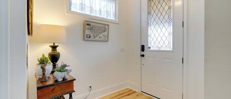A hallway with a wooden floor, a white door with a decorative glass panel, a wall-mounted map, a lamp, and a small table with decorative items.