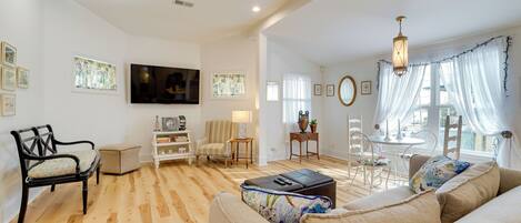 A spacious living room with a wooden floor, a beige sofa, a black armchair, a TV mounted on the wall, and a dining area with a table and chairs.