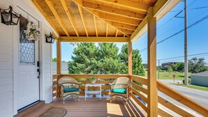 Une véranda en bois avec deux fauteuils en osier et une petite table, donnant sur une rue bordée d’arbres et de bâtiments.