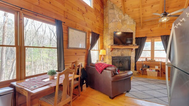 Un interior de cabaña acogedor con paredes de madera, una chimenea de piedra, un televisor y un comedor con mesa y sillas.