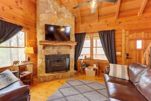 A cozy cabin interior with a stone fireplace, leather sofas, a flat-screen TV, and a wooden ceiling.