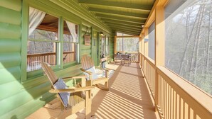 Un porche de madera con revestimiento verde, barandillas de madera y vista a árboles. Hay dos mecedoras y una mesa con sillas.