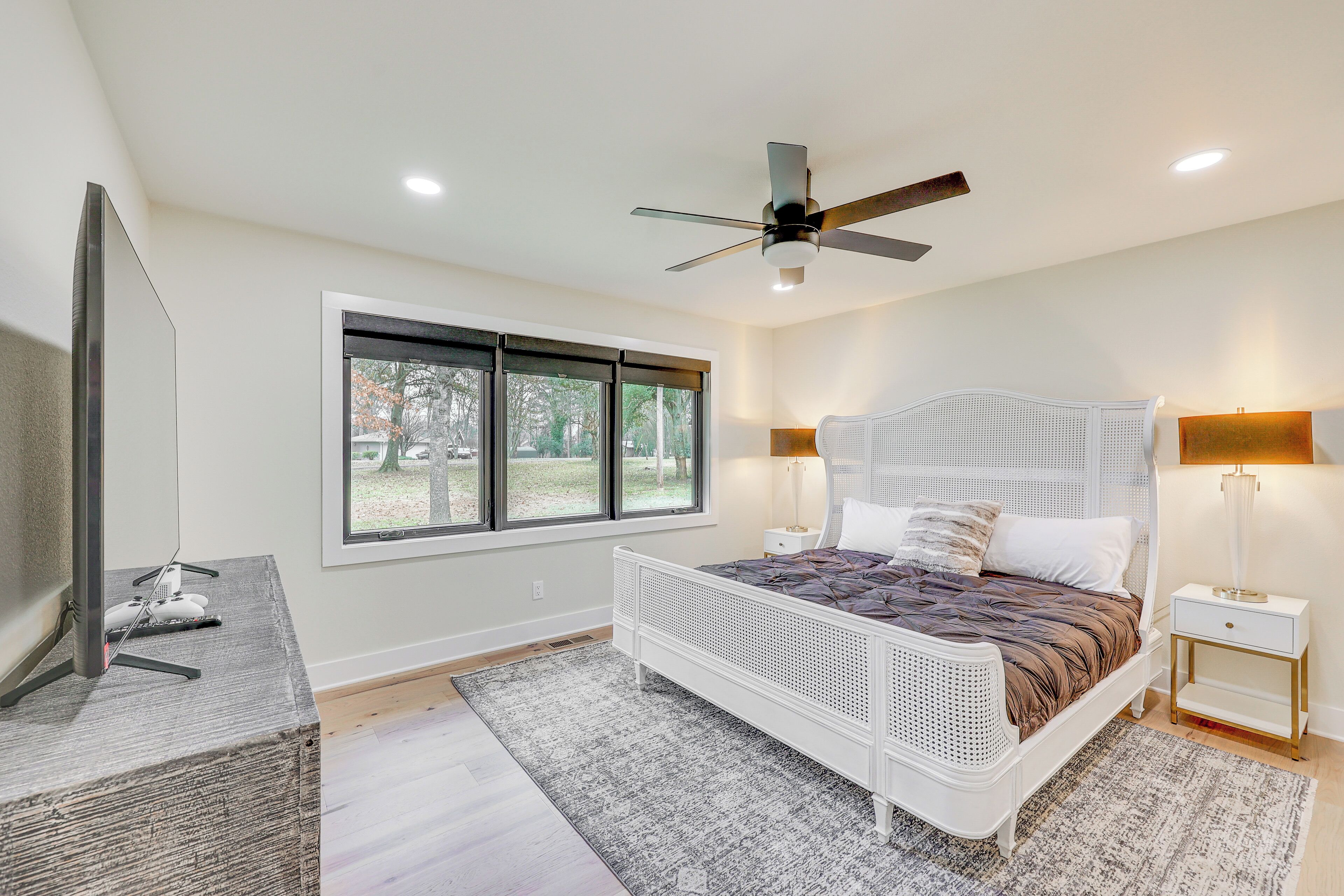 A bedroom with a bed, a TV, a ceiling fan, a window, and a nightstand with a lamp.