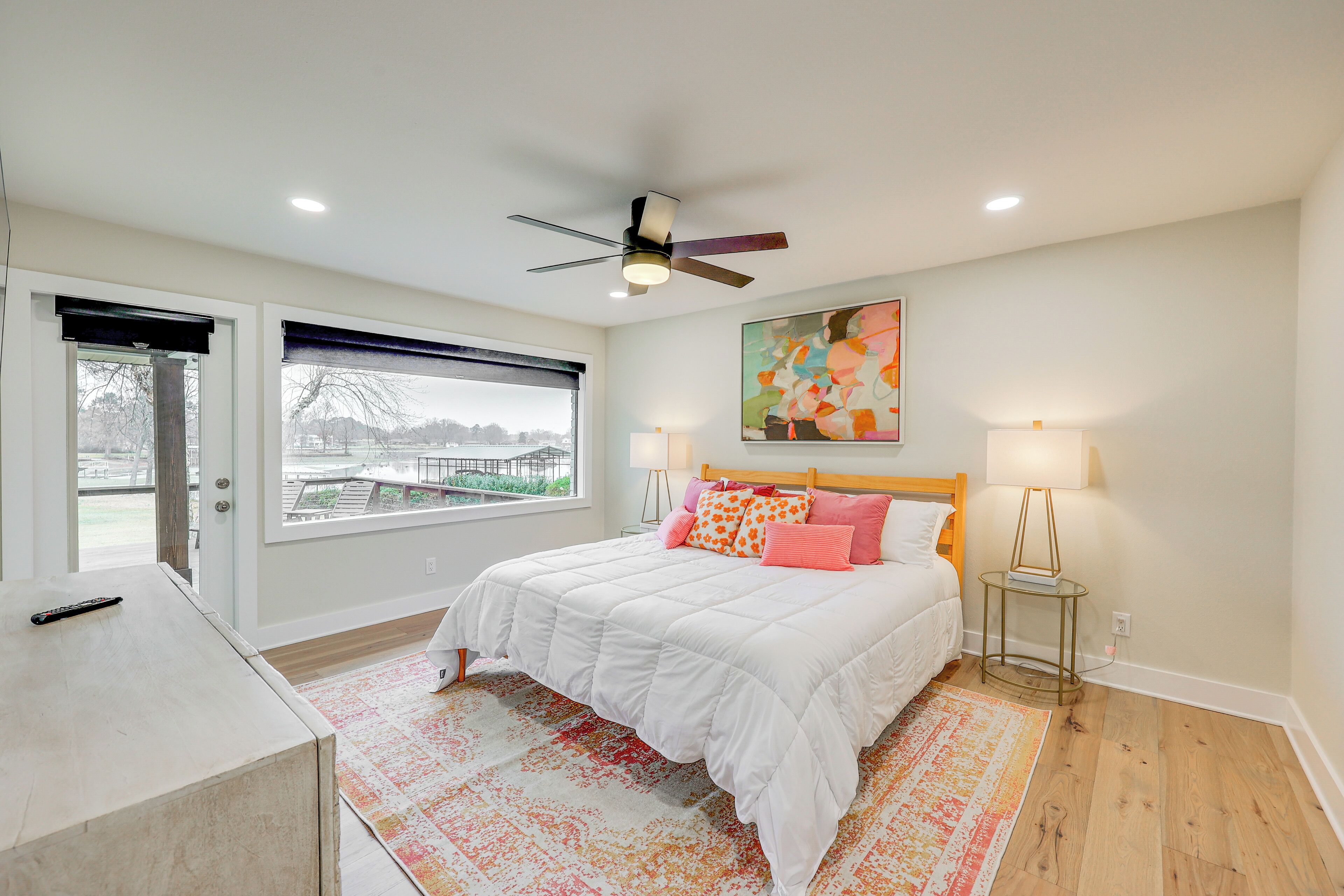 A bedroom with a bed, a ceiling fan, a window with blinds, a painting on the wall, and a bedside table with a lamp.
