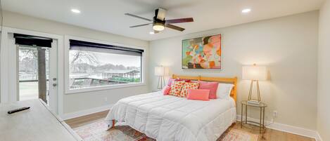 A bedroom with a bed, a ceiling fan, a window with blinds, a painting on the wall, and a bedside table with a lamp.