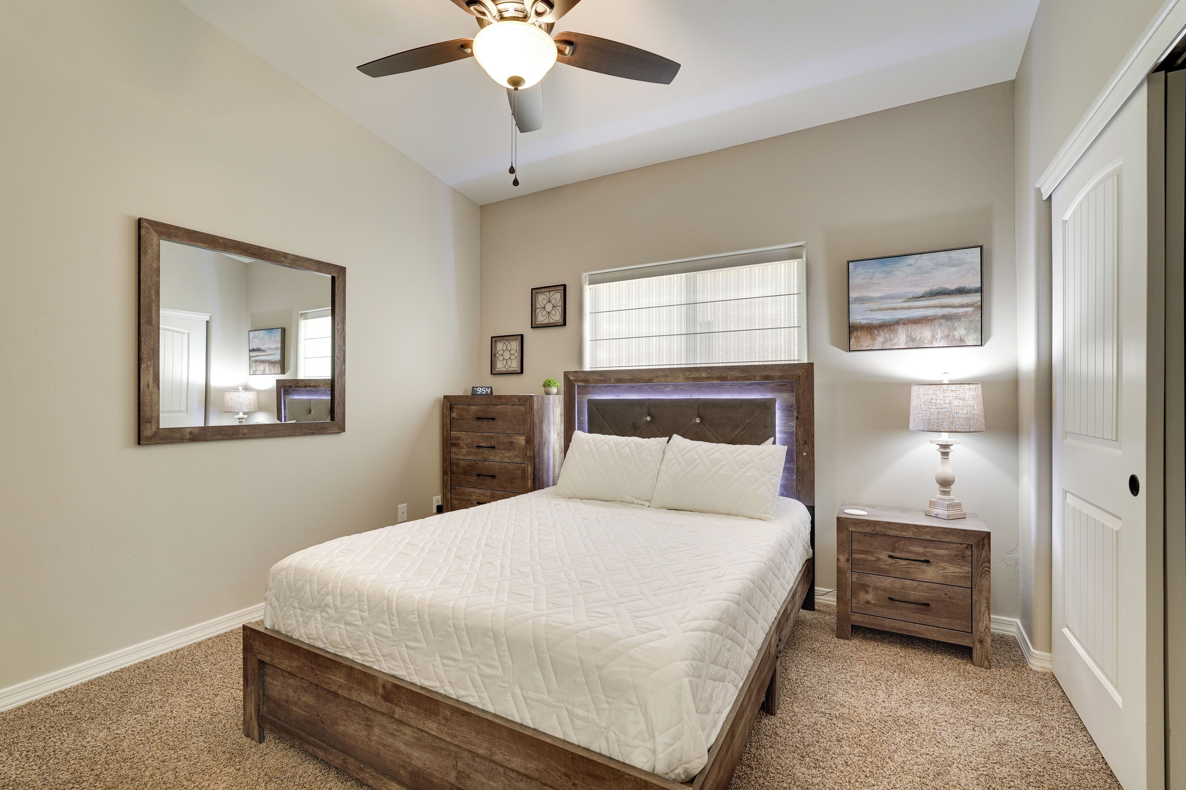 A bedroom with a bed, a nightstand, a ceiling fan, a mirror, and a window with blinds.
