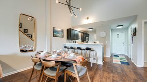 A dining area with a wooden table set for four, a kitchen in the background, and a hallway leading to a door.