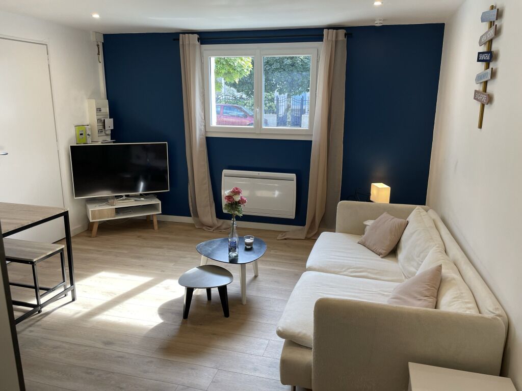 A modern living room with a beige sofa, a small round table with a vase, a flat-screen TV, and a radiator.