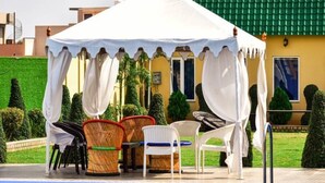 A covered outdoor seating area with chairs and a table, surrounded by a well-maintained garden and a yellow building with a green roof. - SPARSH CANOPY RESORT (Jaipur)