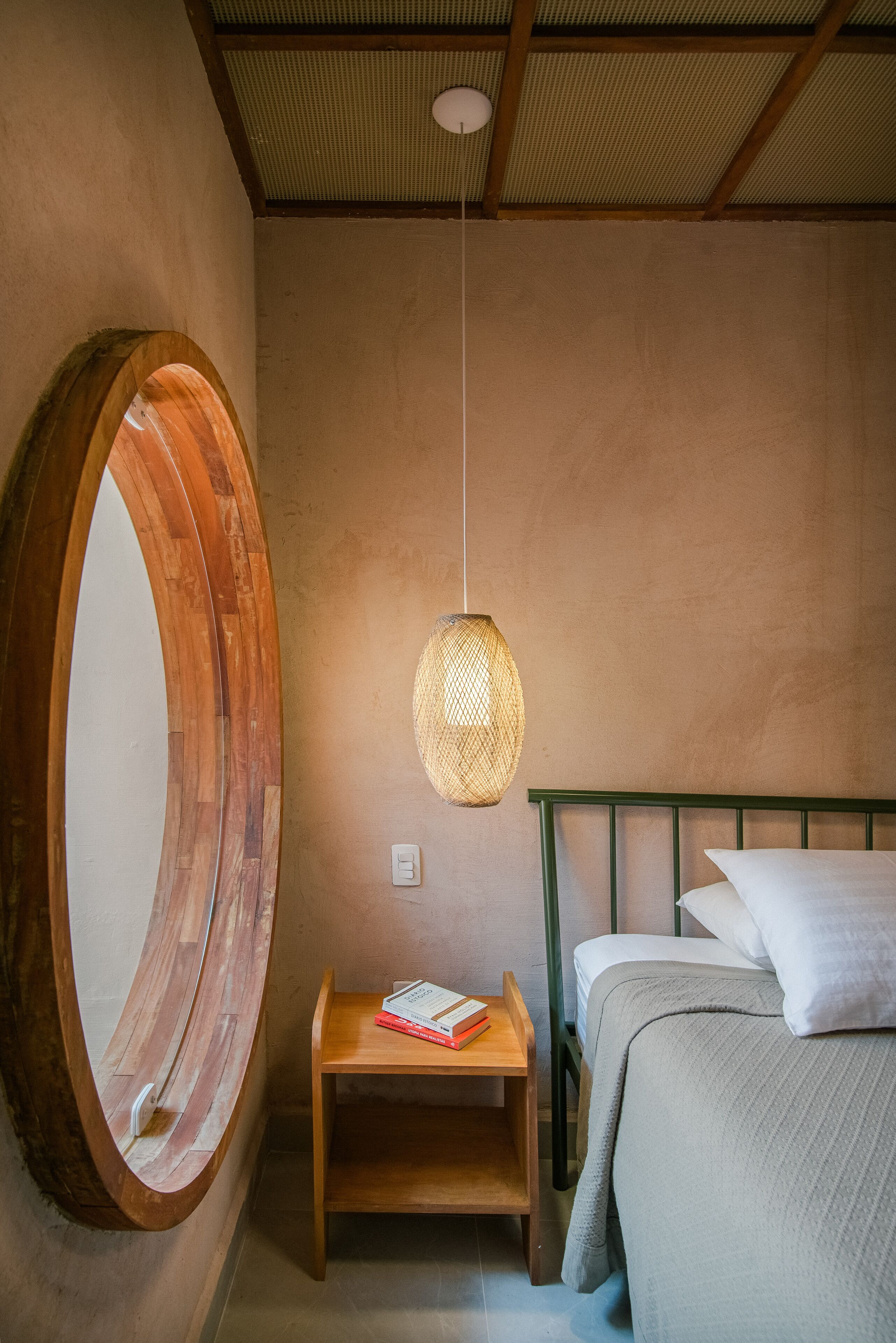 Quarto com espelho redondo, mesa de cabeceira com livros, luminária pendente e cama com almofadas.