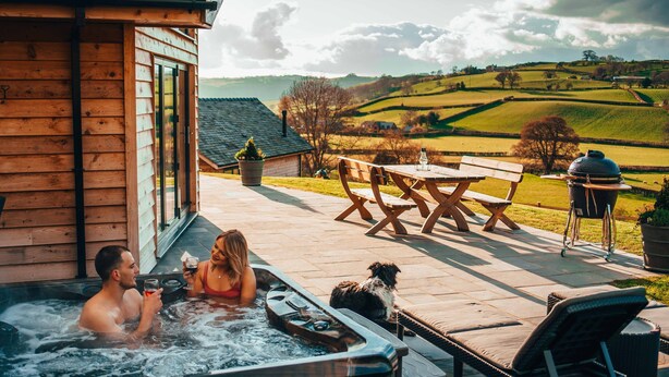 A wooden cabin with a hot tub, outdoor seating, and a scenic view of rolling hills.