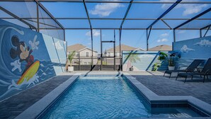 An indoor swimming pool with a Mickey Mouse mural, lounge chairs, and a view of houses through a glass enclosure. - 3725pcl-windsor Island Resort 5 Bedroom Townhouse by RedAwning (Davenport)