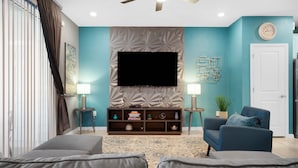 A living room with a flat-screen TV mounted on a textured wall, a blue armchair, a sofa, and a patterned rug. - 3725pcl-windsor Island Resort 5 Bedroom Townhouse by RedAwning (Davenport)