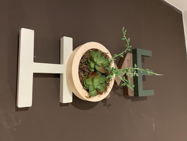 Ein hölzerner Pflanzkasten mit einer Sukkulente und einem kleinen Zweig, platziert zwischen den Buchstaben „H“ und „O“ des Wortes „HOME“ an einer braunen Wand.