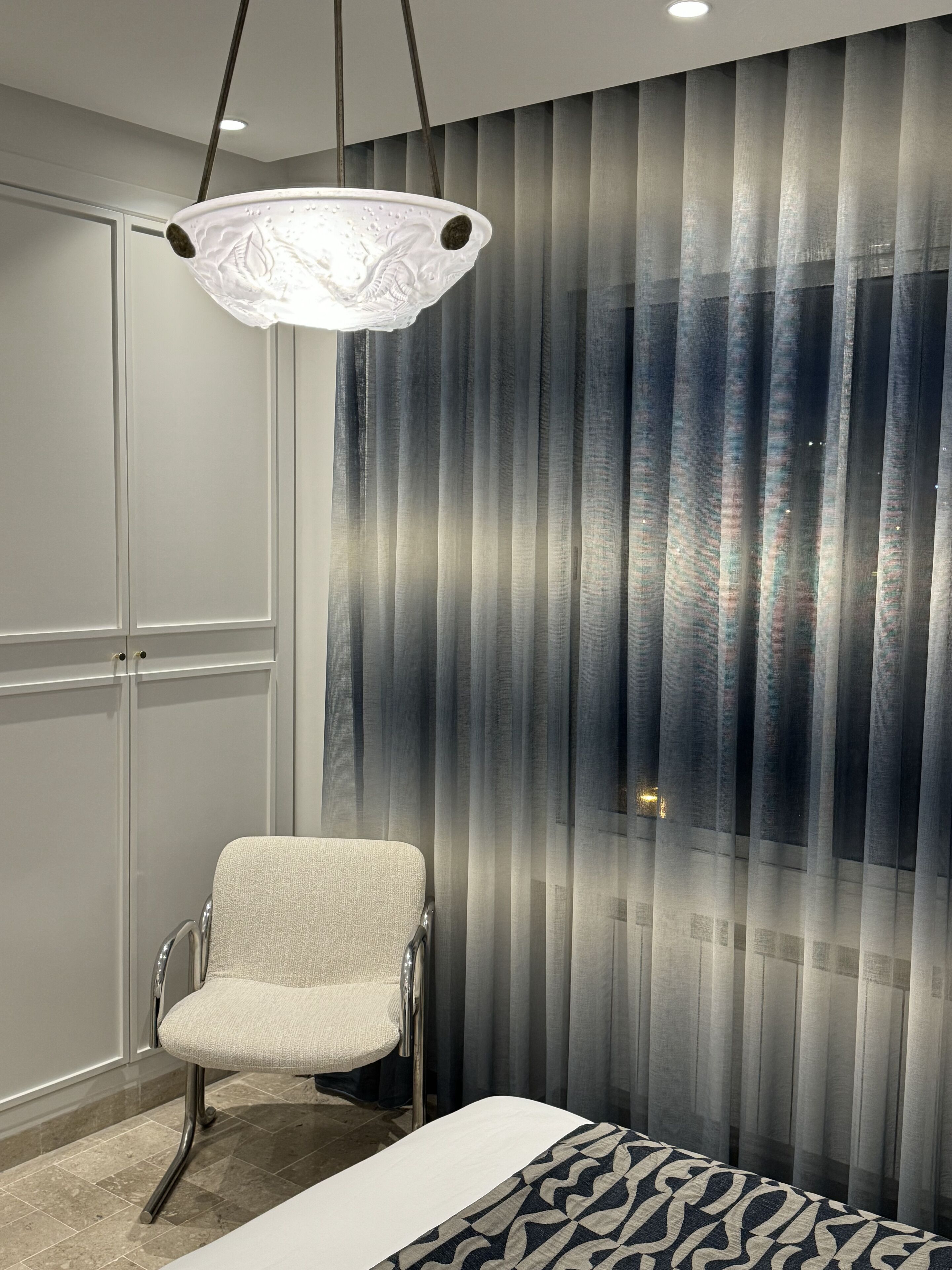 A modern hotel room with a bed, a chair, a large window with vertical blinds, and a hanging light fixture.