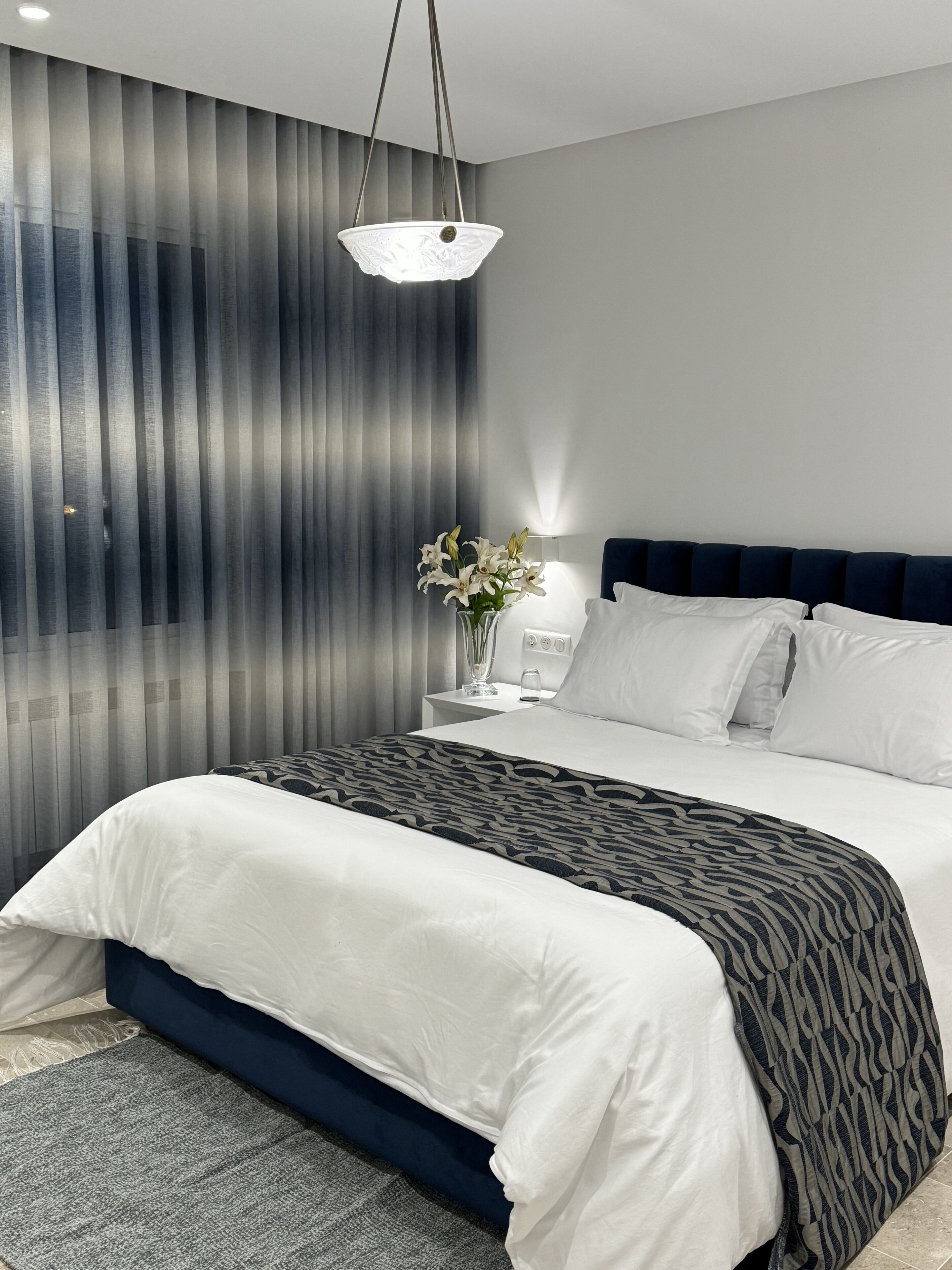 A modern bedroom with a bed, a nightstand, a vase with flowers, and a hanging light fixture.