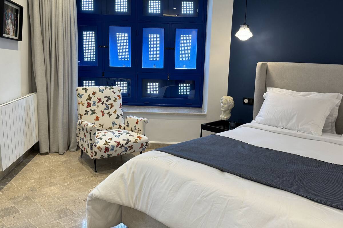 Une chambre à coucher comprenant un lit, une chaise, une fenêtre avec des panneaux bleus et un luminaire fixé au mur.
