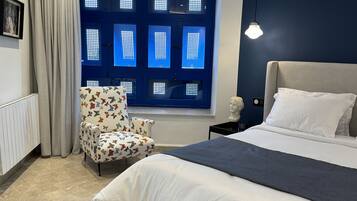 Un dormitorio con cama, una silla, un ventanal con paneles azules y una lámpara fijada a la pared.