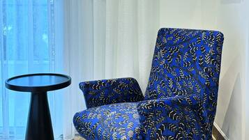 A modern room with a blue patterned armchair, a small round table, and a view of a building through sheer curtains.