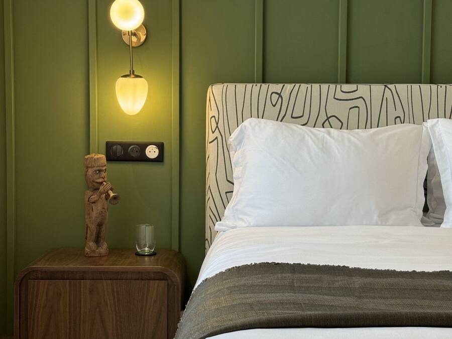 A bedroom with a bed, a nightstand, a wall-mounted lamp, and a small wooden sculpture.
