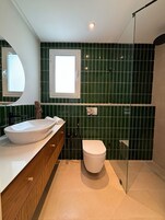Un baño con lavamanos blanco, tocador de madera, inodoro blanco y zona de ducha.