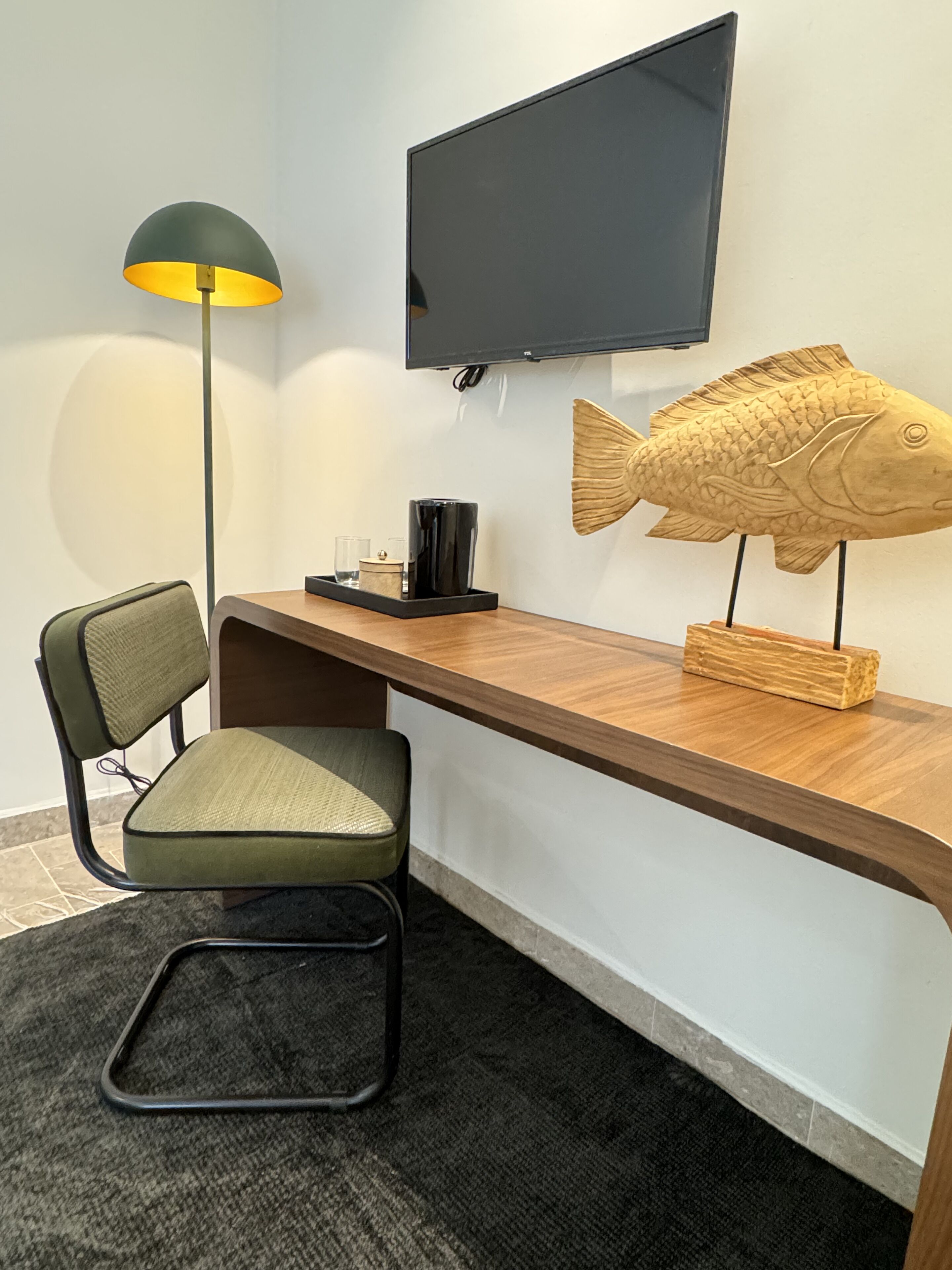 A modern room with a wooden desk, a green chair, a floor lamp, a mounted TV, and a decorative fish sculpture.
