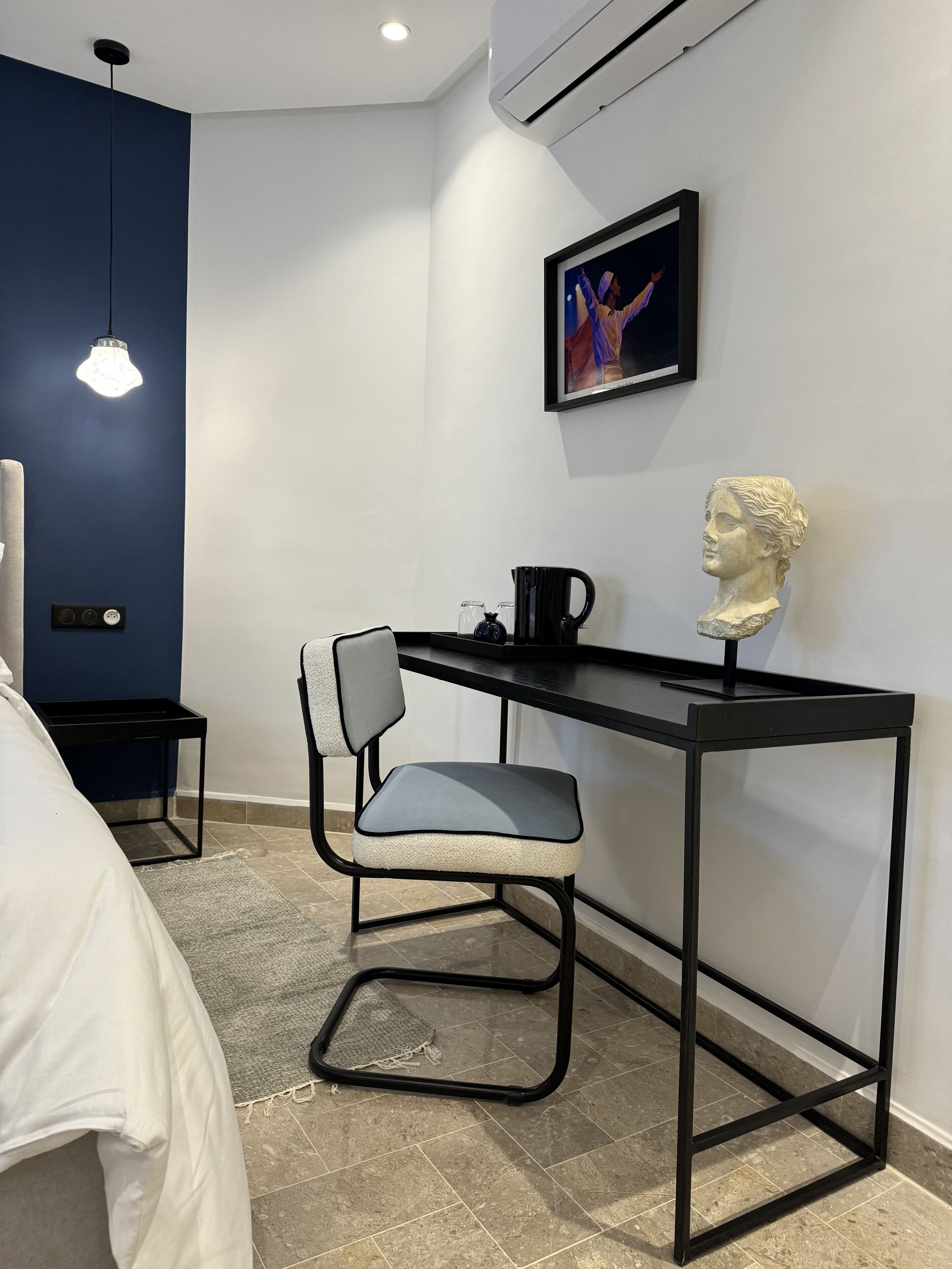 Una habitación moderna con una cama, un escritorio, una silla, una lámpara colgante, un cuadro enmarcado y una escultura de busto.