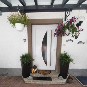 A white door with a frosted glass panel, flanked by two black planters with green plants, a small figurine, and a decorative doormat. - Großzügige 2-zimmer Wohnung mit Eigenem Garten (Sulz am Neckar)
