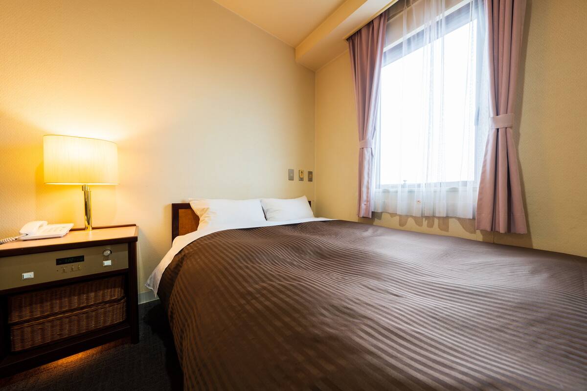 A hotel room with a bed, a nightstand with a lamp, and a window with curtains.