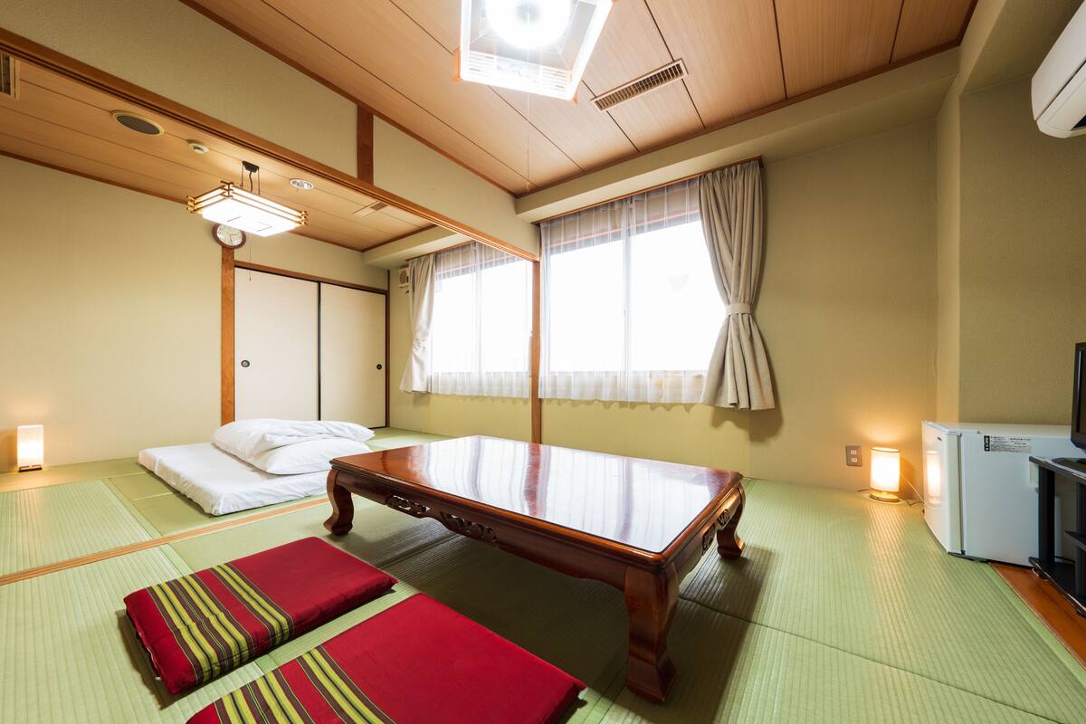 A traditional Japanese room with tatami flooring, a low wooden table, a futon bed, and a small refrigerator.