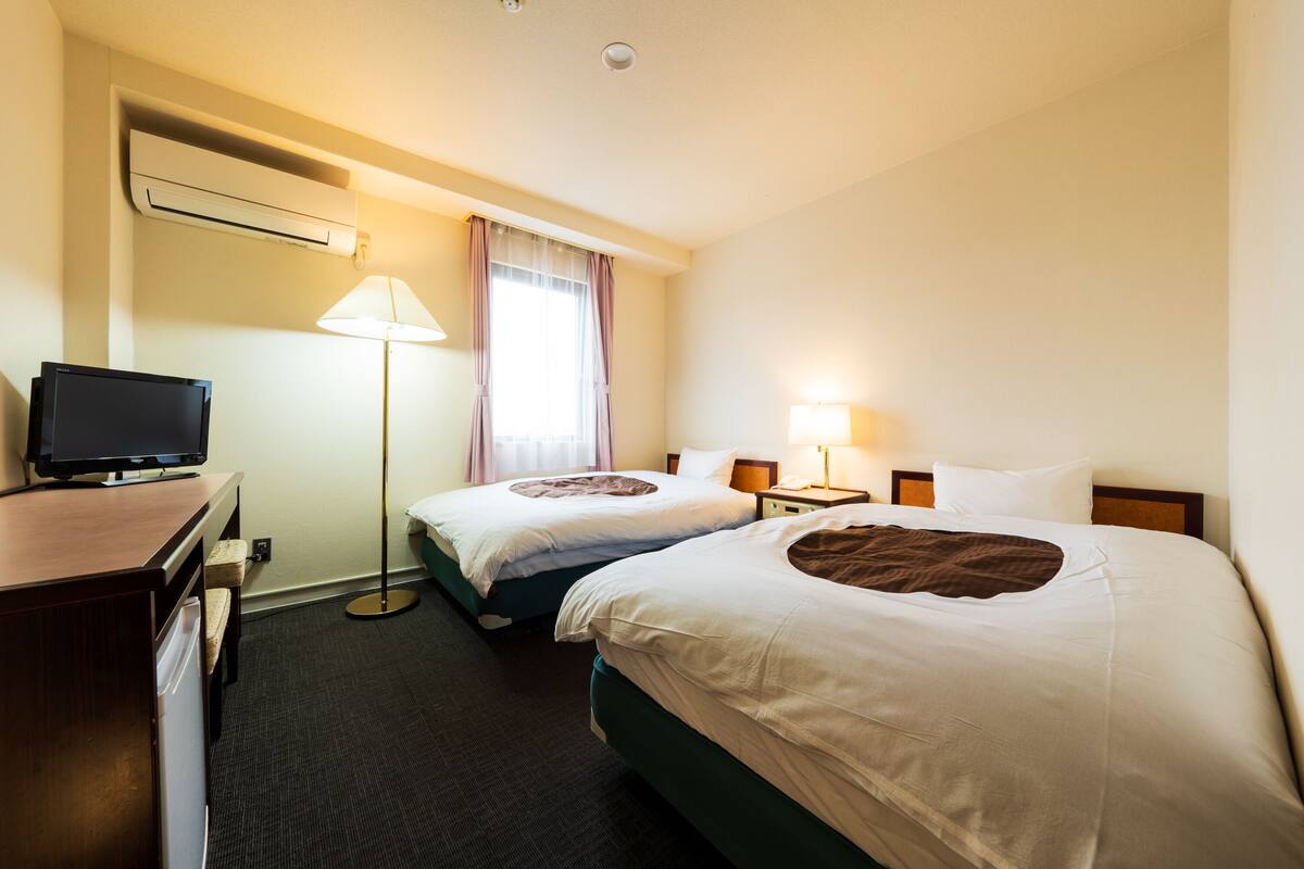 A hotel room with two beds, a desk with a television, a lamp, and an air conditioning unit.