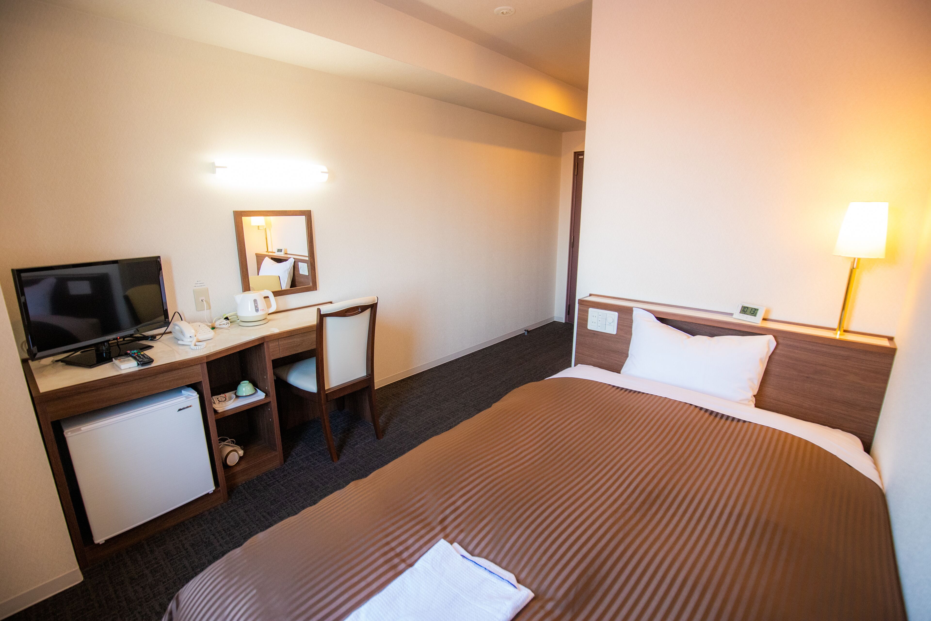 A hotel room with a bed, a desk with a television, a chair, a small refrigerator, and a lamp.