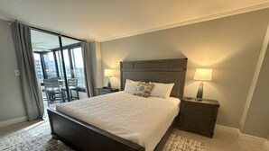 A hotel room with a large bed, two bedside tables with lamps, a view of the city through a sliding door, and a balcony with chairs.