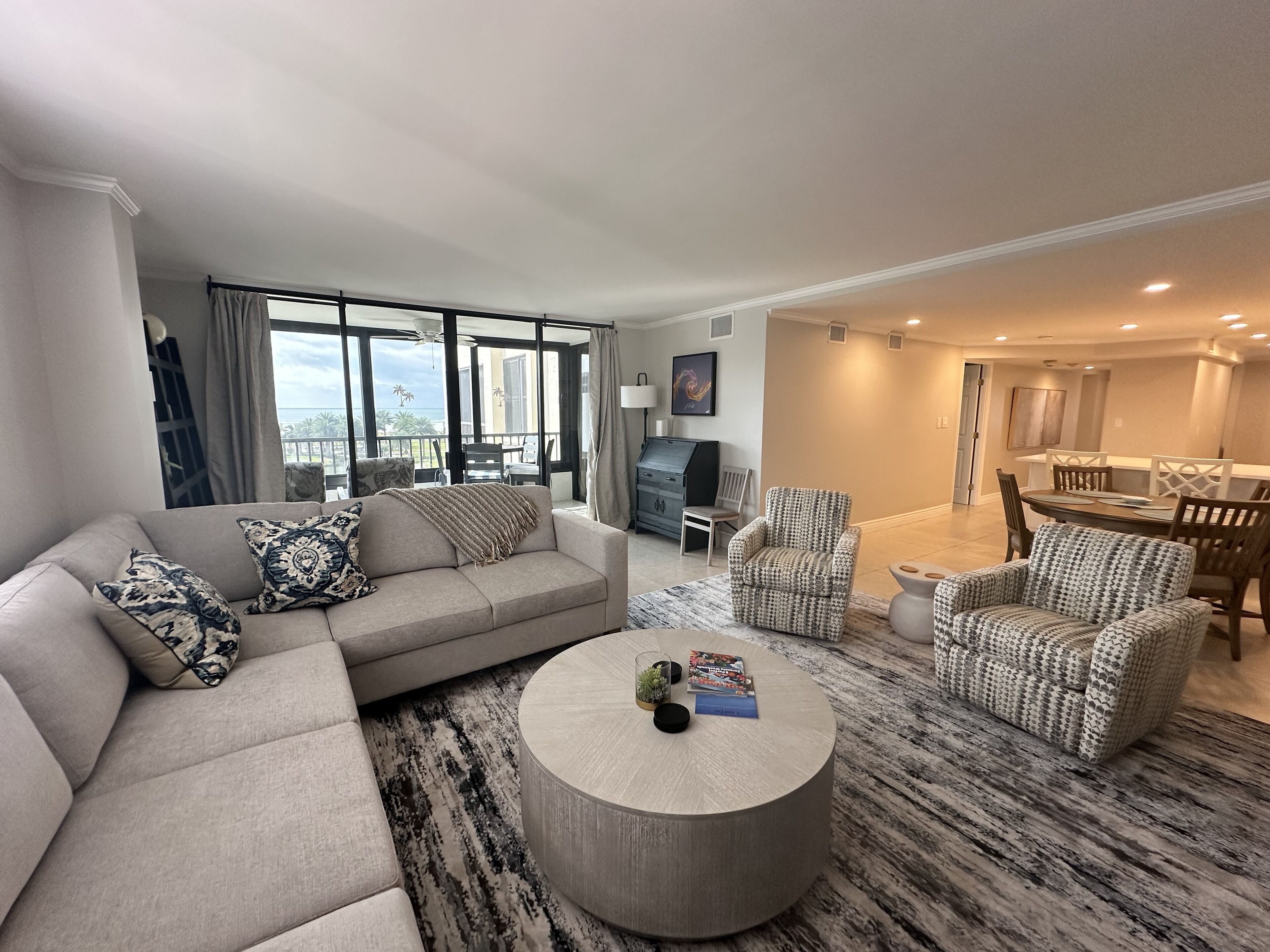 A modern living room with a sectional sofa, patterned armchairs, a round coffee table, and a dining area with a table and chairs.