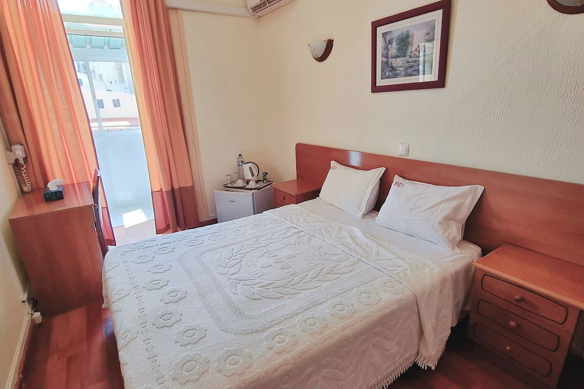 A hotel room with a bed, bedside tables, a nightstand, a wall-mounted air conditioner, a framed picture, and a window with curtains.