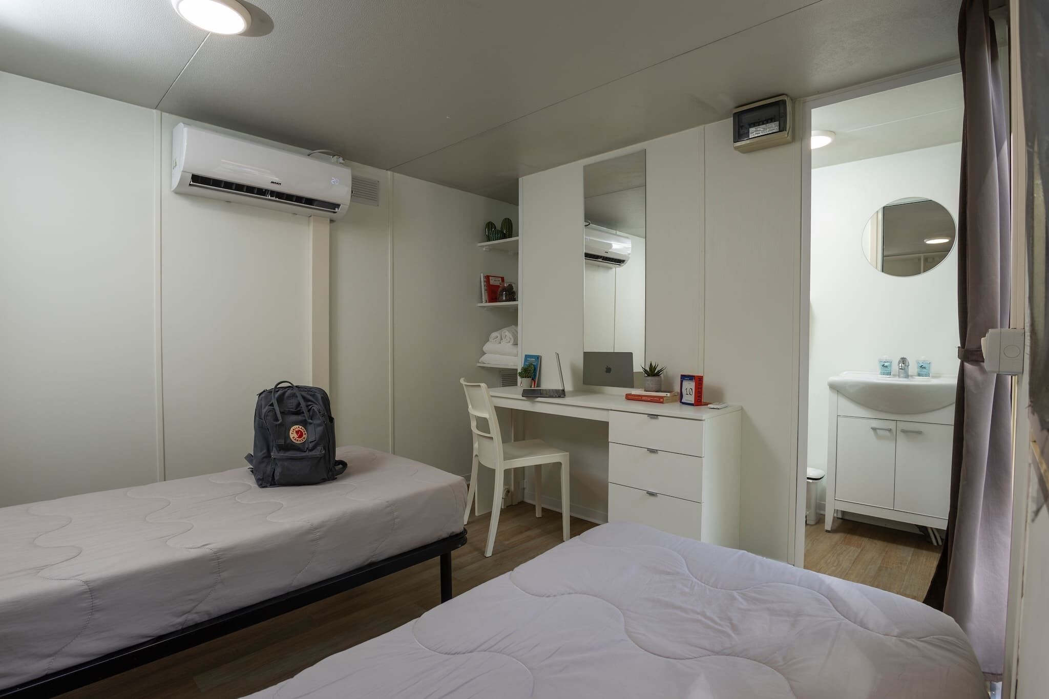 A hotel room with two single beds, a desk with a chair, a wardrobe, a bathroom with a sink and mirror, and an air conditioning unit.