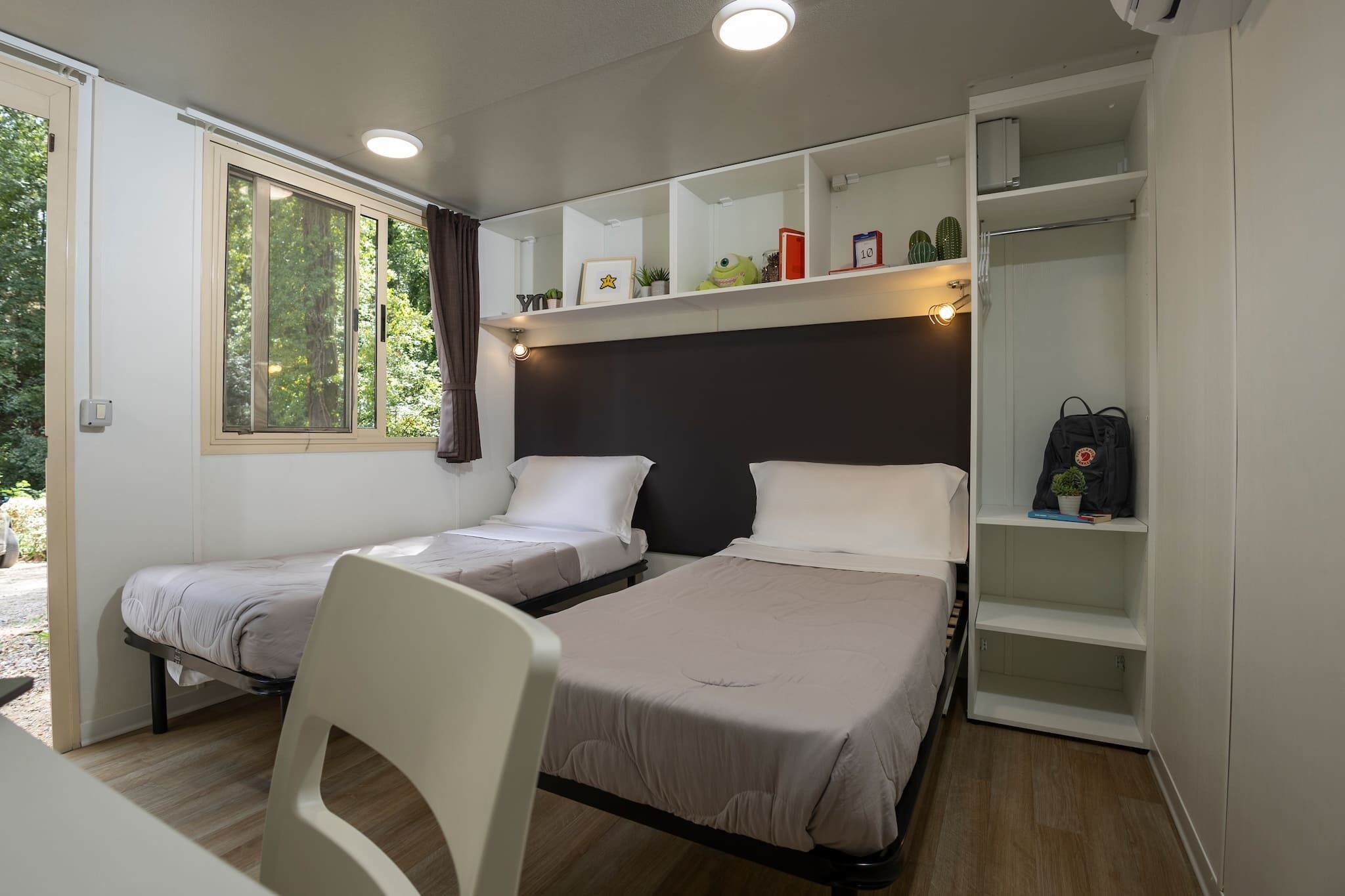 A compact room with two beds, a desk, and a chair. There is a window with curtains, a shelf with various items, and a backpack on the floor.