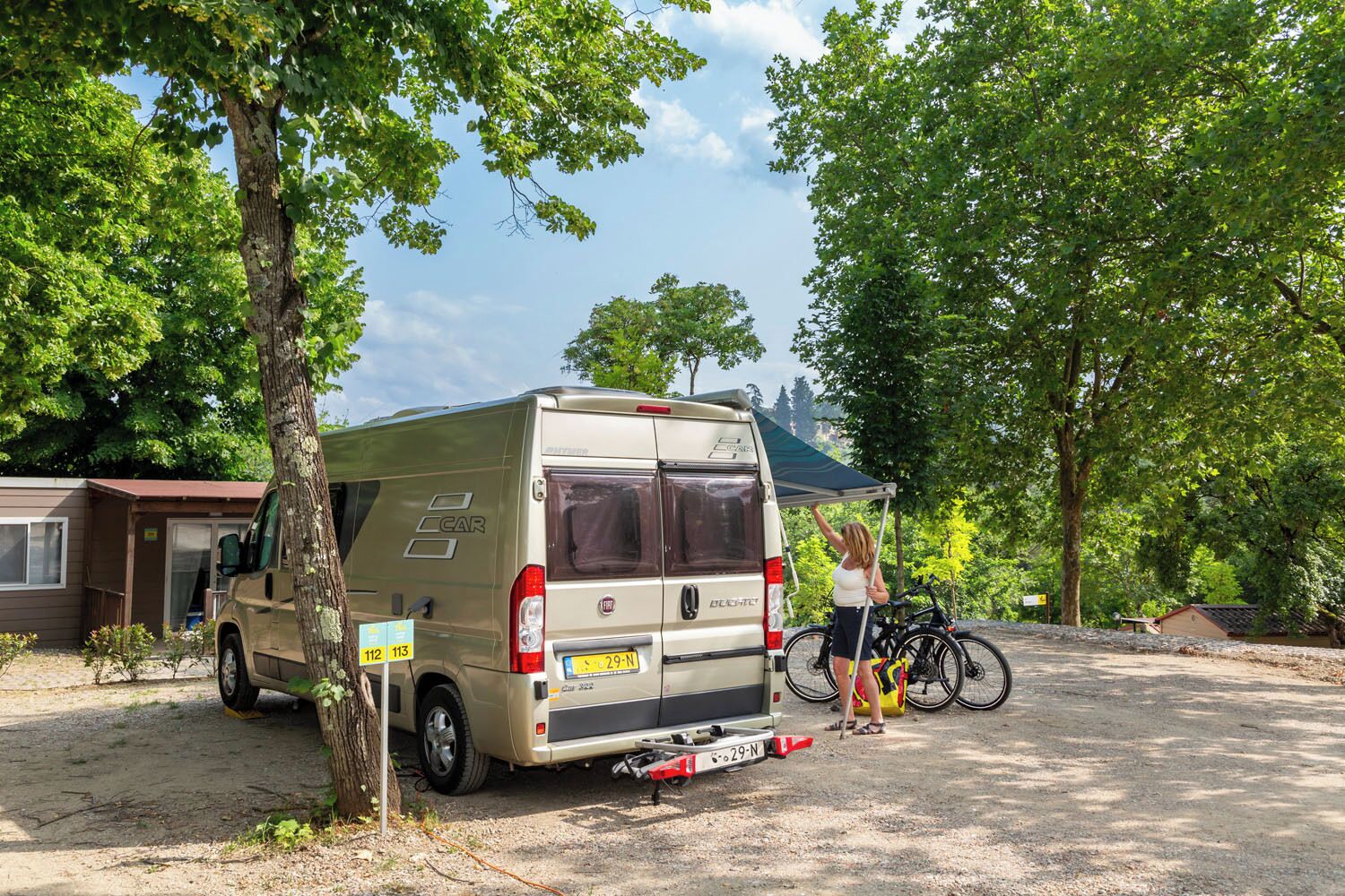 A beige van parked in a campsite with trees and a small building in the background.