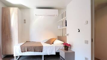 Un dormitorio con cama, una mesita de noche, un armario, aire acondicionado instalado en la pared y una estantería con libros.