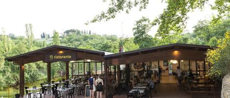 An outdoor restaurant with tables and chairs, a sign reading "ristorante", and a view of trees and hills in the background.