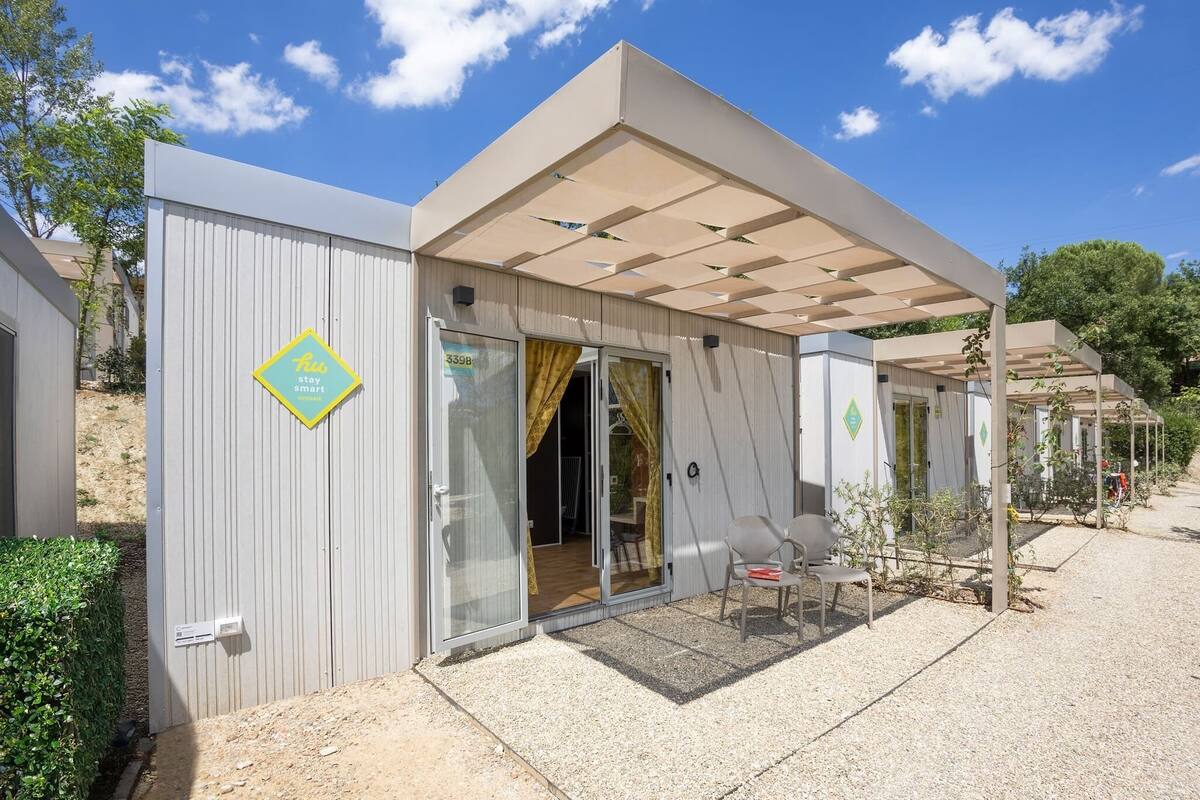 Een modern, gelijkvloers gebouw met een overdekte patio, een glazen schuifdeur als ingang en een klein buitenzitgedeelte met een tafel en stoelen.