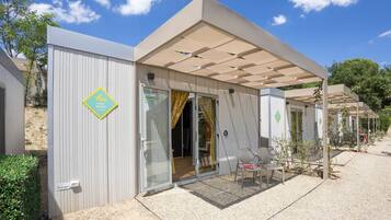 Een modern, gelijkvloers gebouw met een overdekte patio, een glazen schuifdeur als ingang en een klein buitenzitgedeelte met een tafel en stoelen.
