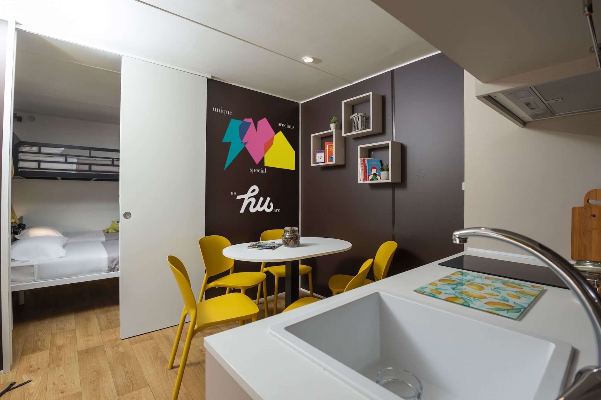 Quarto compacto com beliche, uma mesa redonda pequena com cadeiras amarelas, pia branca e parede com design colorido.