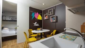 Quarto compacto com beliche, uma mesa redonda pequena com cadeiras amarelas, pia branca e parede com design colorido.
