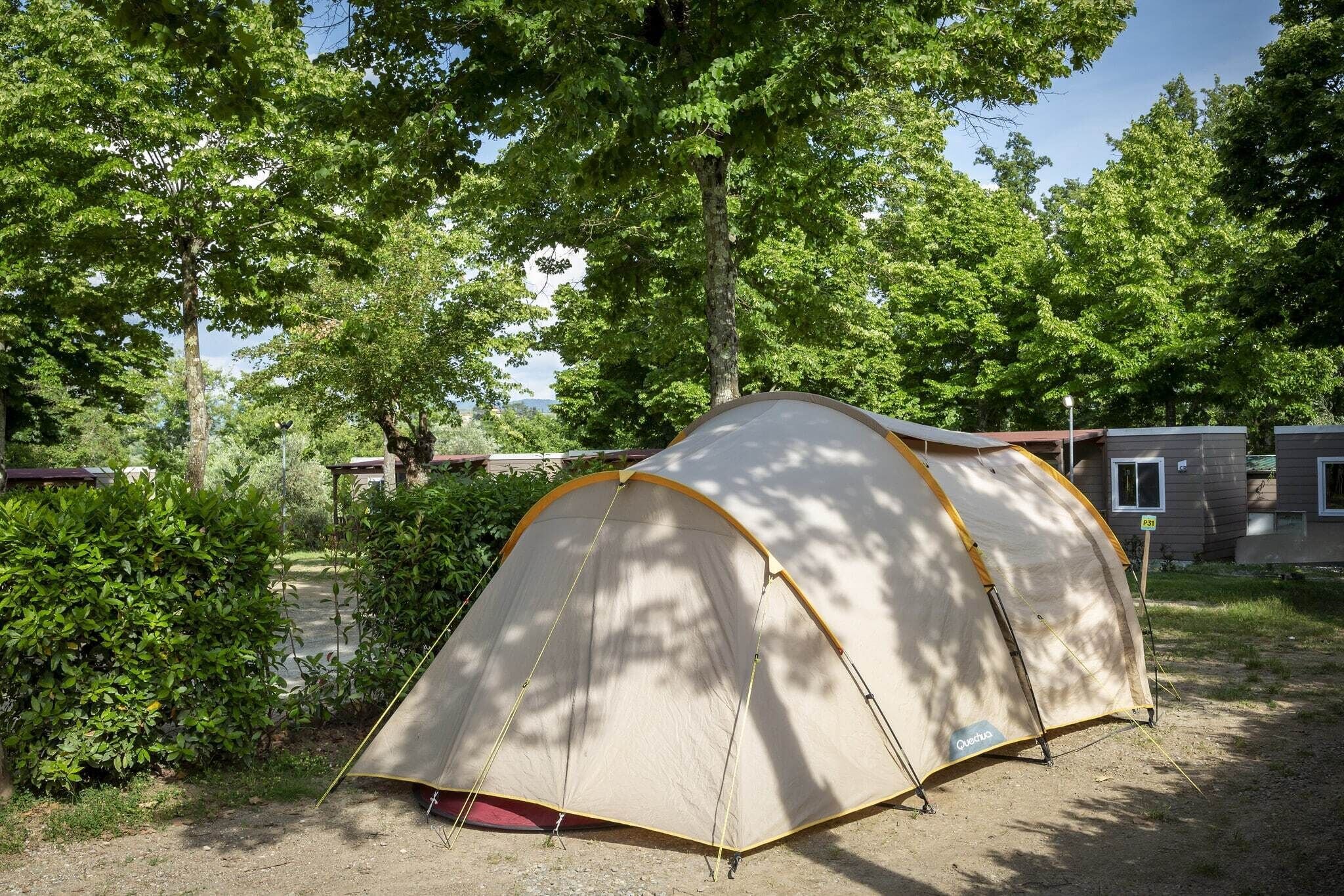 A dome tent with a beige and yellow color scheme, set up in a natural, wooded area with trees and a building in the background.