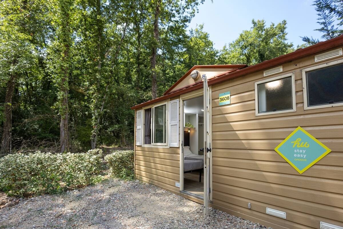 Een kleine, eenvoudige houten hut met een openstaande deur, waar een bed en een raam zichtbaar zijn. Op de deur staat een bord met '360B' en op de muur staat 'hius stay easy OPENAIR'.