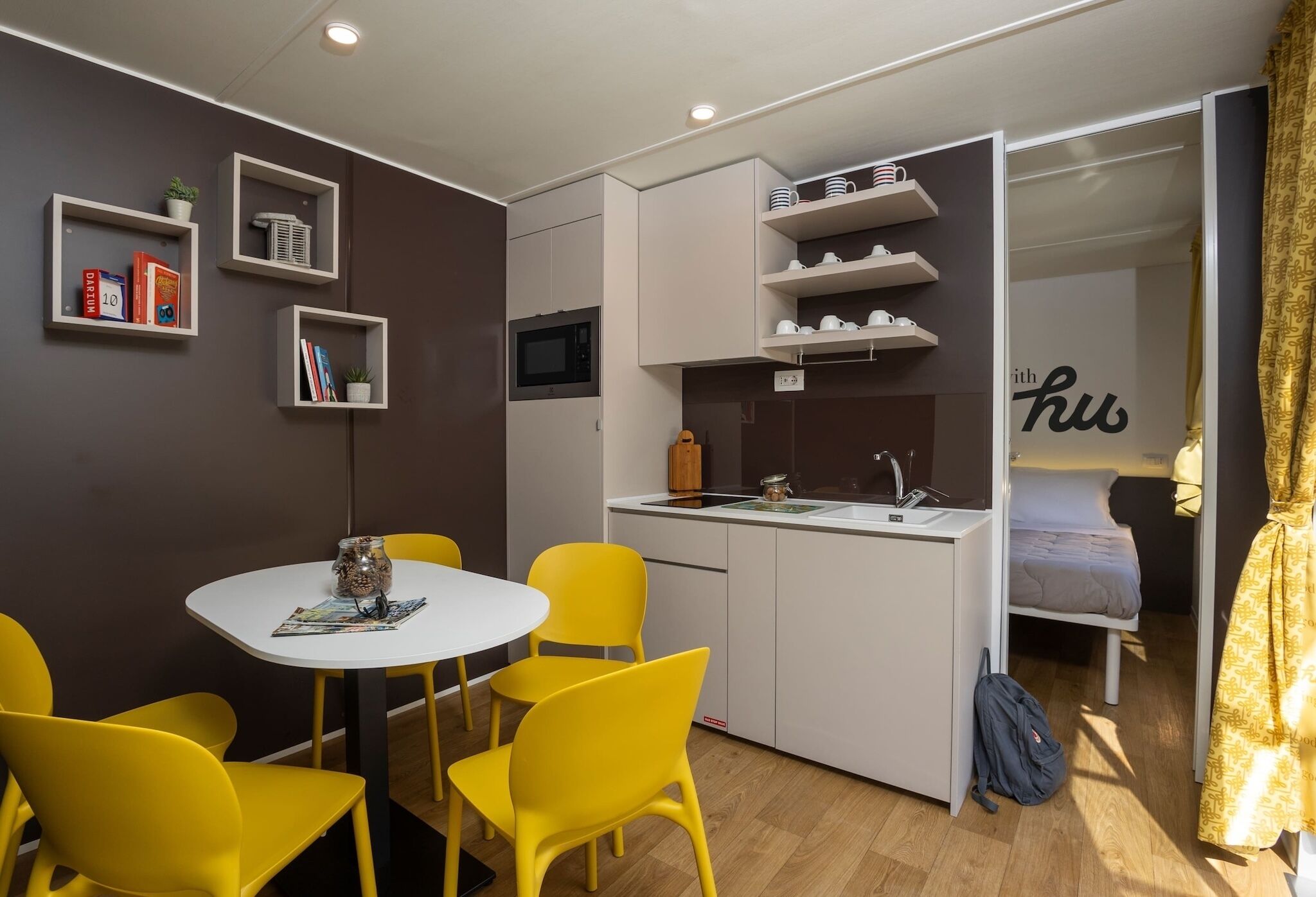 A compact room with a kitchenette, a dining area with yellow chairs, a bed, and a small shelf with books and a plant.