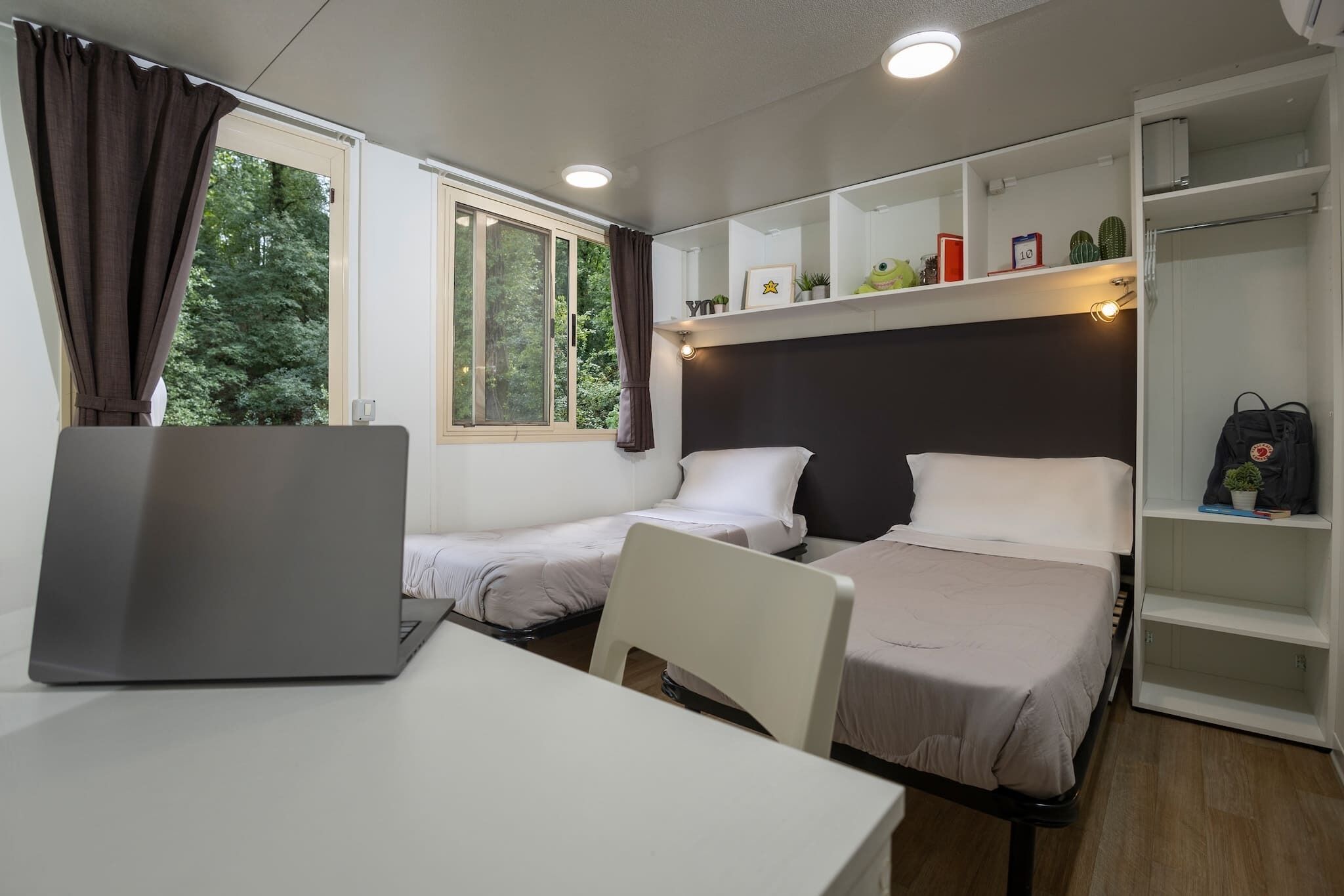 Una habitación pequeña con dos camas, un escritorio y un ordenador portátil. Hay una ventana con cortinas, una estantería con adornos y un armario.