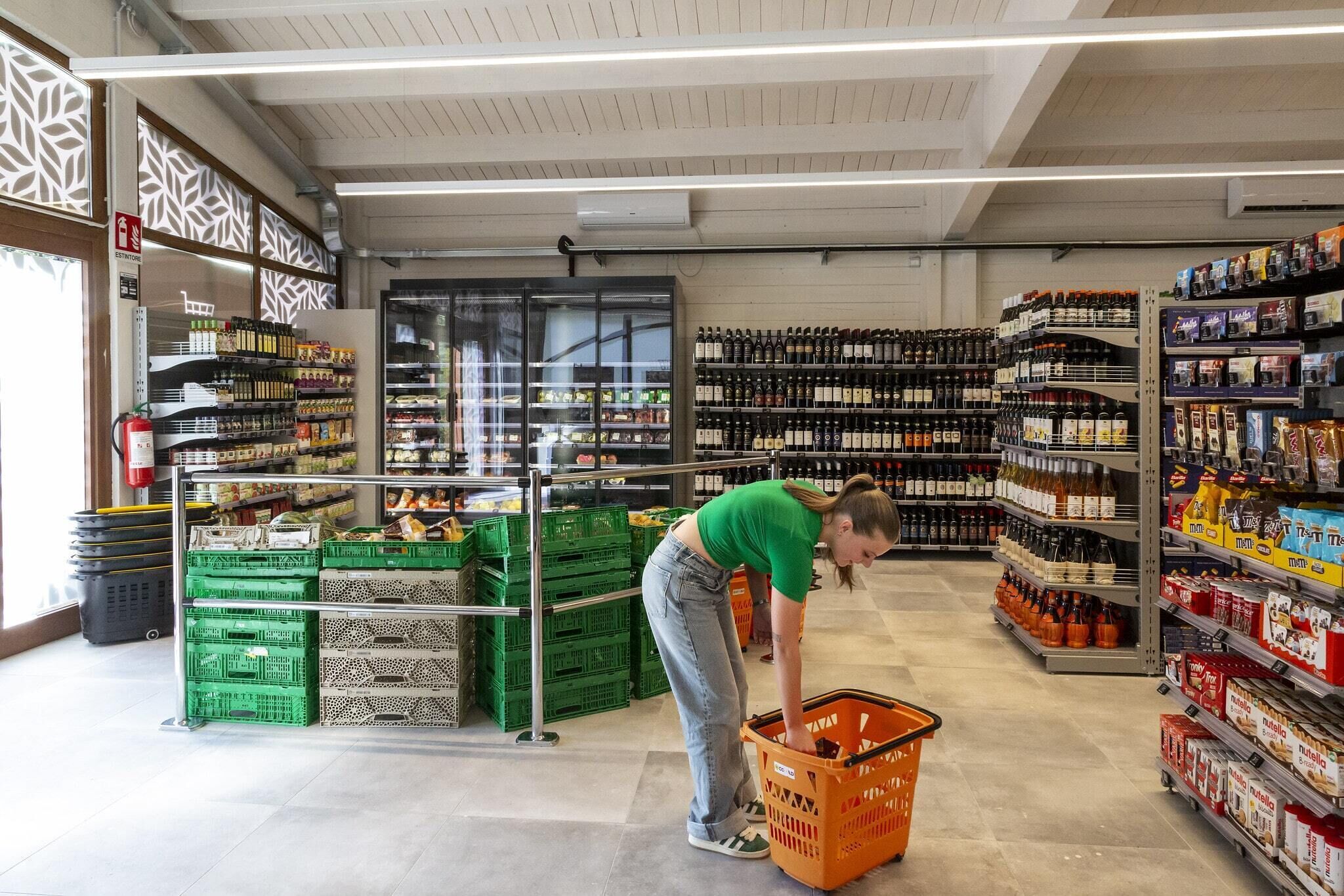 A grocery store aisle with shelves stocked with various products, a refrigerated section, and a person bending over a shopping basket.